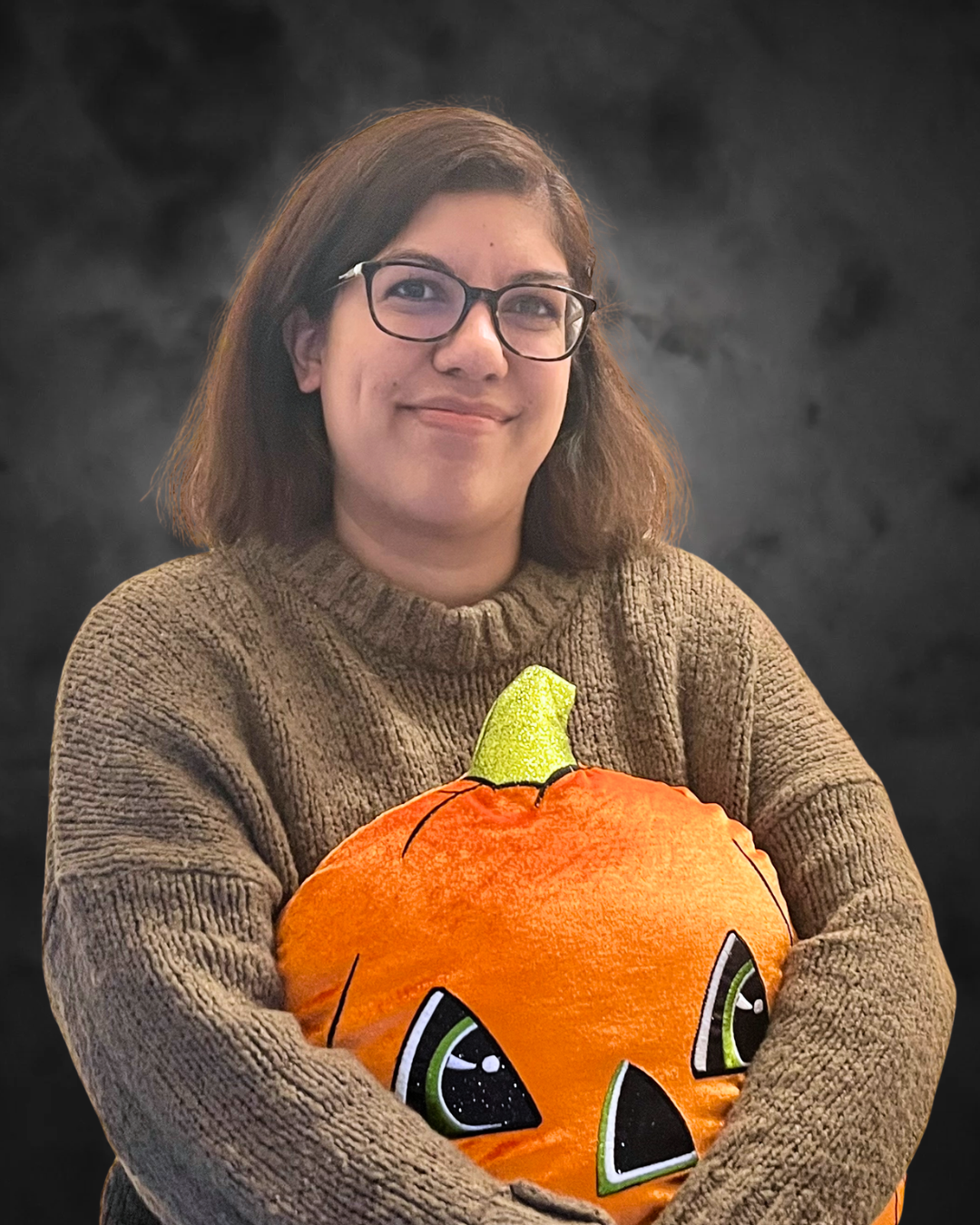 Alana Sookoor, Registered Social Worker and Psychotherapist at Elite Counselling, smiles while holding an orange pumpkin plushie against a dark background.