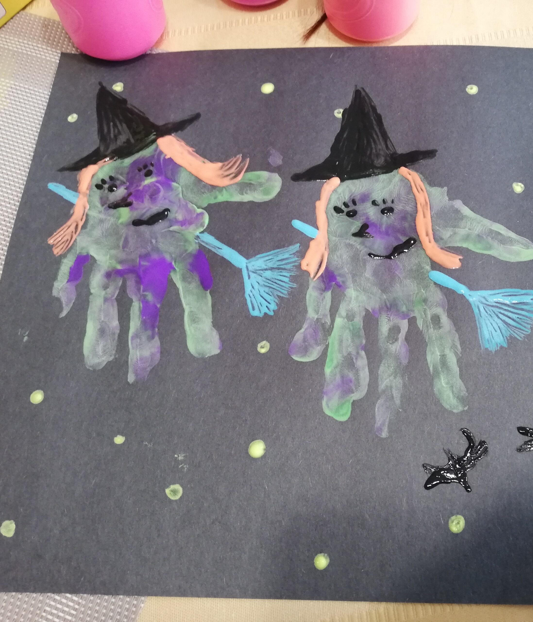 Two colourful Halloween witches made from green and purple painted handprints on black construction paper, decorated with pointed hats, orange hair, blue broomsticks, and painted faces.