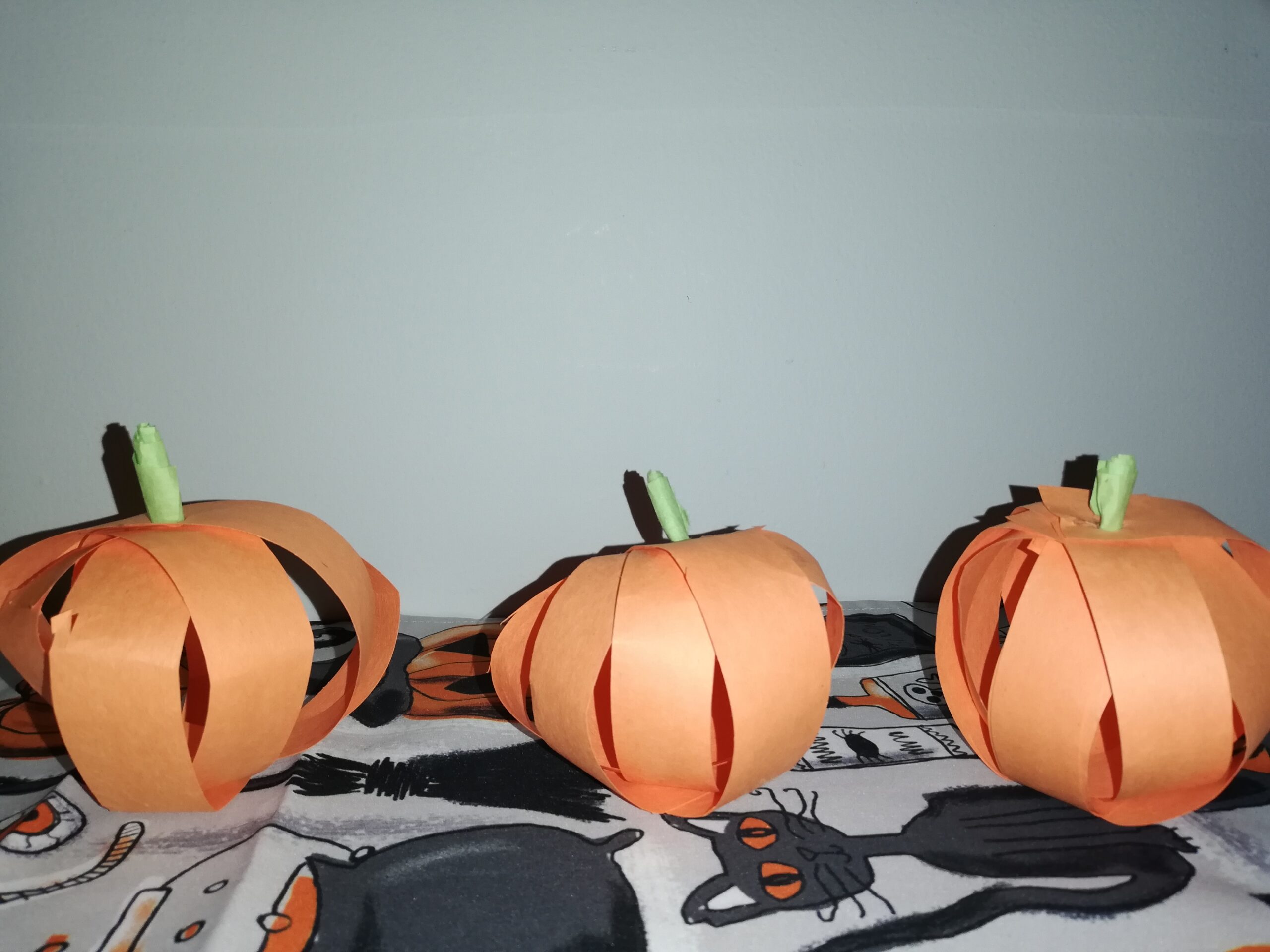 Three handmade orange paper pumpkins with green stems displayed on a Halloween-themed tablecloth featuring black cats, showcasing a simple Halloween craft for children.