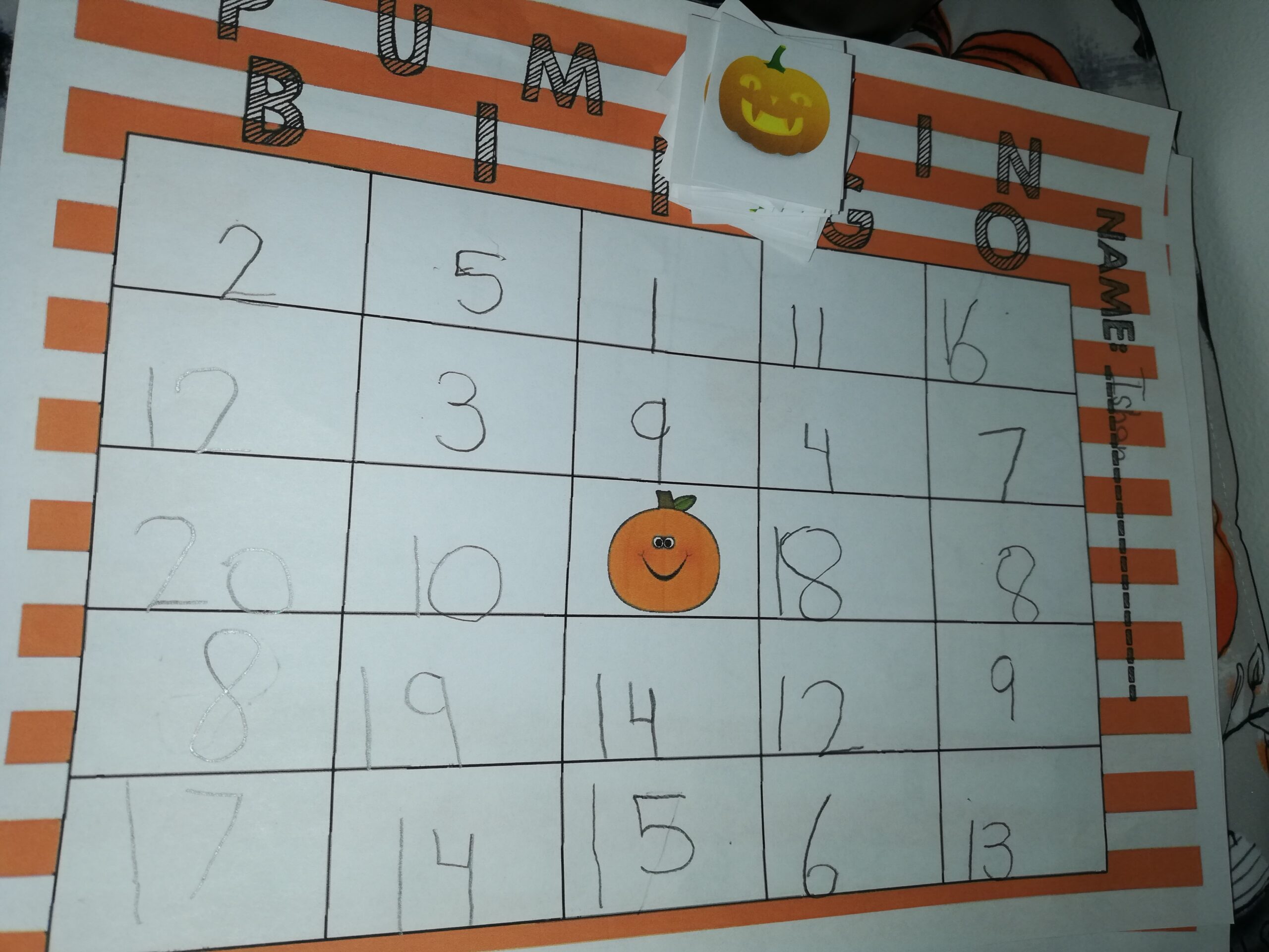 Homemade Halloween bingo game featuring a numbered 5-by-5 grid, a smiling pumpkin in the centre, and pumpkin-themed calling cards for a fun children's learning activity.