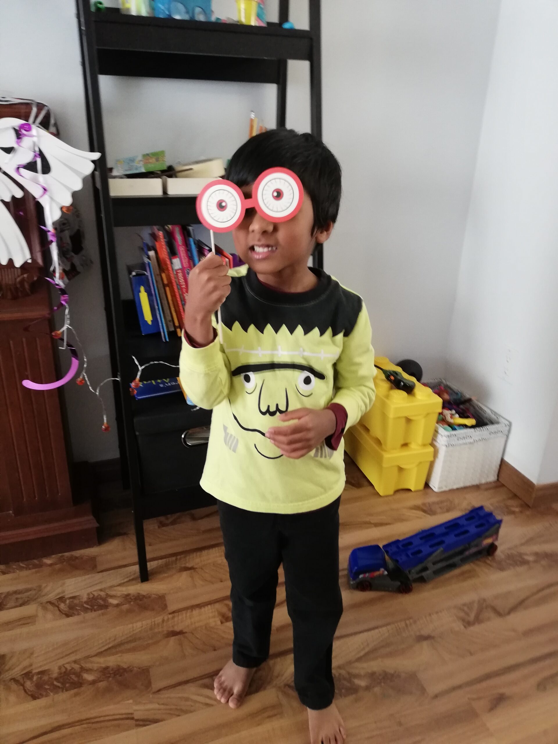 Young child wearing a Frankenstein-themed shirt and holding playful eyeball glasses over their eyes during Halloween pretend play at home.