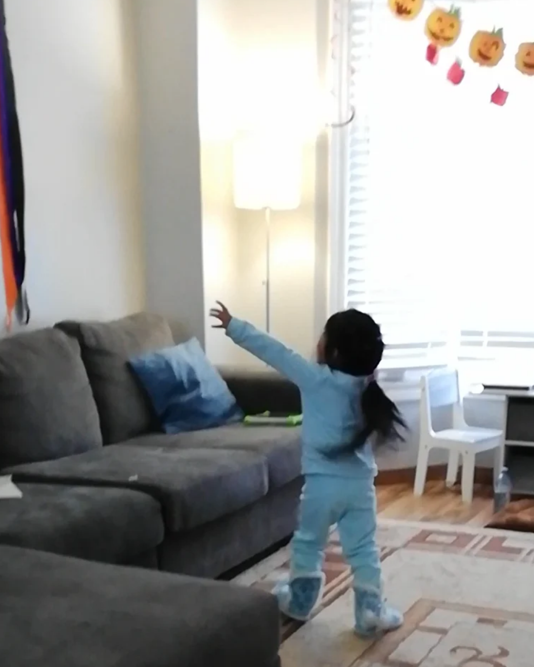 Young child in blue pyjamas dancing with arms outstretched in a living room decorated with Halloween pumpkins.